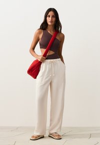 Femme en crop top asymétrique marron et pantalon blanc taille haute à cordon tient un sac à épaule rouge, debout sur un sol carrelé clair.