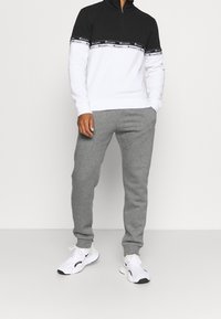 Felpa nera e bianca con zip a metà e banda con logo, abbinata a pantaloni jogger grigi e scarpe sportive bianche. Design semplice e vestibilità confortevole.