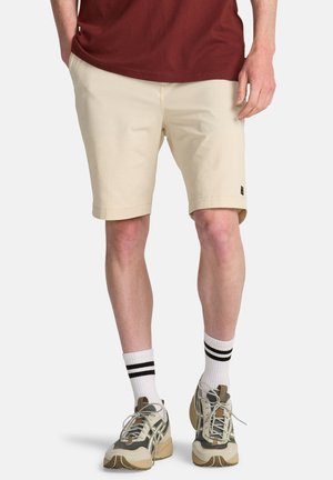 Beżowe spodenki wykonane z bawełny, z płaskim przodem i małą naszywką z logo po lewej stronie. Noszone z bordową koszulą i sportowymi butami.