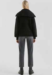 Zwarte, oversized textuurjas met een grote kraag, gedragen met grijze textuurbroek en zwarte enkellaarsjes, gezien vanaf de achterkant.