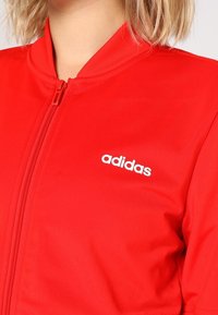 Veste rouge zippée avec col côtelé et logo Adidas blanc sur la poitrine gauche, portée par une personne aux cheveux blonds courts.