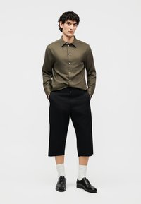 Hombre joven de pie con las manos en los bolsillos, vistiendo una camisa verde oliva abotonada, pantalones negros recortados, calcetines blancos y zapatos de vestir negros.