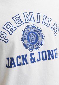 Valkoinen puuvillapaita, jossa on sininen teksti "PREMIUM JACK & JONES" ja ympyrälogo "DENIM ACADEMY" sekä vuosi "SINCE 1990."