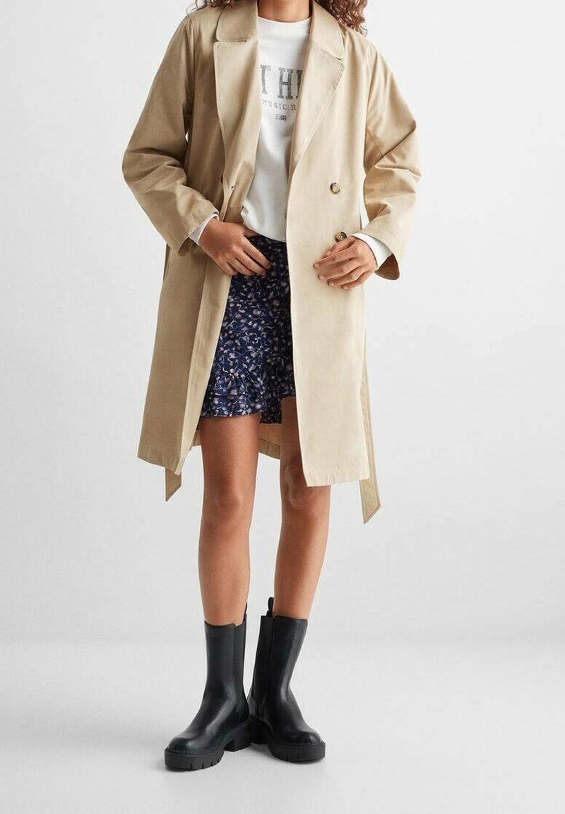 Personne portant un trench beige, un t-shirt blanc à motif graphique, une jupe bleue à motifs et des bottines noires, debout devant un fond uni.