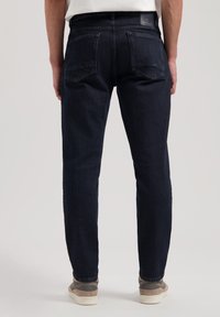 Jeans de mezclilla azul oscuro con un corte recto, dos bolsillos traseros y un acabado texturizado. Llevados con zapatillas grises y beiges.