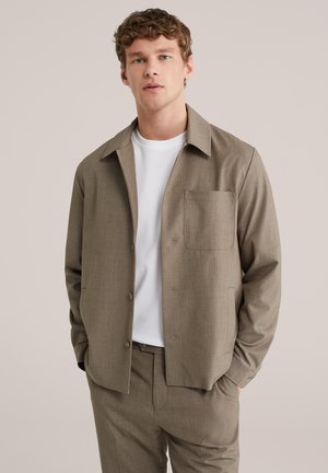 Noor mees kannab beežikat nööbitavat jope suurt rinnataskut, valge t-särgi ja sobivate beežide pükstega, käed taskutes.