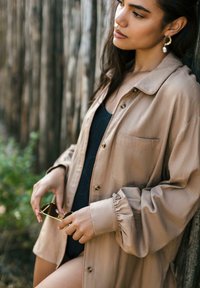 Camicia beige oversize con colletto e chiusura a bottoni; presenta una tasca frontale e polsini arricciati. Gli accessori includono occhiali da sole e orecchini a goccia.