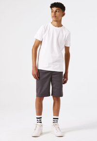 Camiseta blanca de manga corta combinada con pantalones cortos grises. El atuendo incluye zapatillas altas blancas y calcetines a rayas en blanco y negro con logos.