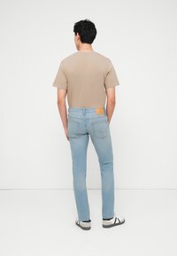 Άνδρας που στέκεται με την πλάτη, φοράει ανοιχτόχρωμο μπλε τζιν, μπεζ κοντομάνικο πουκάμισο και λευκά αθλητικά παπούτσια με μαύρες λεπτομέρειες, μπροστά σε ένα απλό λευκό φόντο.