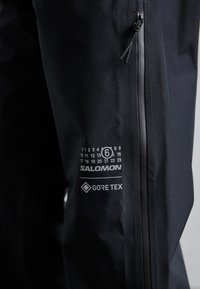 Fekete vízálló nadrág sima textúrával, oldalra zippzárral, nyomtatott naptár részlettel és Salomon és Gore-Tex márkajelzéssel.