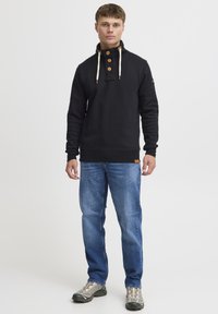 Suéter negro con cuello alto, tres botones de madera y cordones de crema; combinado con jeans azules y zapatillas de senderismo grises.