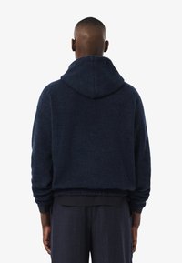 Uomo visto di spalle che indossa una felpa con cappuccio di un blu scuro e pantaloni coordinati su uno sfondo bianco uniforme.