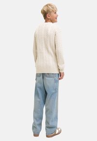 Ein blondes Kind trägt einen cremefarbenen Kabelstrickpullover, hellblaue lässige Jeans mit einer Seitentasche und beige Schuhe. Es steht seitlich und lächelt.
