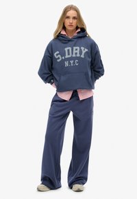 Joven mujer vestida con sudadera navy blue S.DRY NYC, camiseta rosa debajo, pantalones anchos navy a juego y zapatillas beige, de pie frente a un fondo blanco.