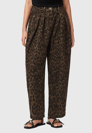 Kvinde iført løstsiddende, højttaljede brune leopardmønstrede bukser kombineret med sorte flade sandaler med stropper mod en ensartet baggrund.