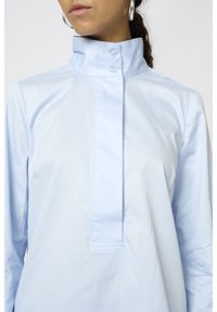 Femme portant une chemise bleu clair à col montant avec une patte de boutonnage devant et de grandes boucles d'oreilles argentées en anneau, montrée des épaules au menton.