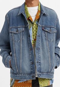 Giacca di jeans con due tasche sul petto, colore azzurro chiaro, chiusura anteriore con bottoni e dettagli in cucitura a contrasto. Indossata sopra una camicia a fantasia.