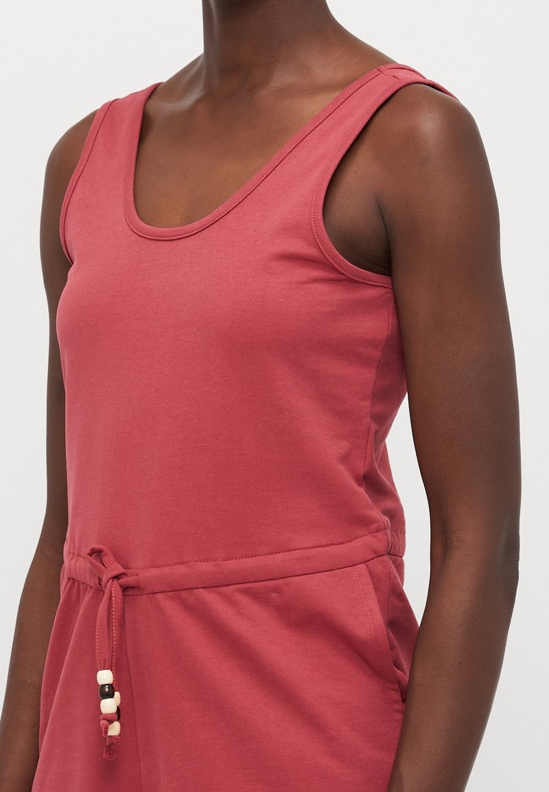 Buste d'une personne portant une robe sans manches rose rougeâtre avec un encolure dégagée et une taille nouée ornée de perles noires et blanches.