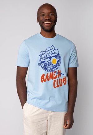 Helles blaues T-Shirt mit einem grafischen Design von Ramen in einer Schüssel mit Essstäbchen und einer Sonne, akzentuiert durch bold roten Text "RAMEN CLUB."