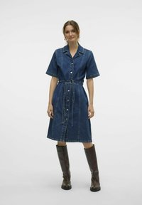 Denimskjortklänning i mörkblått med korta ärmar, figurnära midja, knäppning med knappar och matchande bälte; kombinerad med knähöga bruna stövlar.