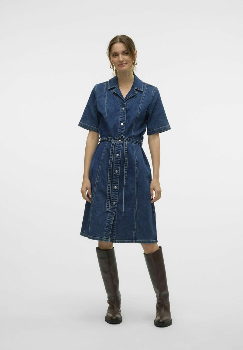 Denimskjortklänning i mörkblått med korta ärmar, figurnära midja, knäppning med knappar och matchande bälte; kombinerad med knähöga bruna stövlar.