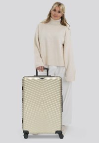 Frau mit blonden Haaren, die einen beigen Rollkragensweater und weiße Hosen trägt, steht und hält einen großen goldenen Hartschalenkoffer mit Rollen.