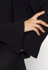 Pull noir en tricot avec manches larges texturées et détails côtelés. Le tissu semble doux avec une coupe ample, mettant l'accent sur le confort.