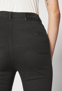 Primer plano de una persona que lleva unos vaqueros negros de Emporio Armani con una parche de la marca y el logotipo de un águila en el bolsillo trasero, combinados con una camiseta blanca.