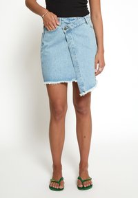 Gonna a portafoglio in denim blu chiaro con orlo sfilacciato, dotata di chiusura frontale con bottoni e tasca laterale. Indossata con infradito verdi.