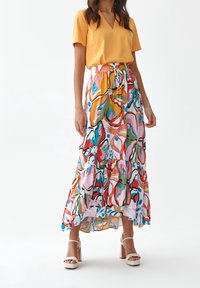 Žena v žlutém krátkém rukávu, barevné květinové maxi sukni s páskem na stažení a bílých vysokých podpatcích na bílém pozadí.