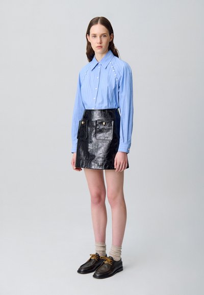Camisa a rayas azul claro con detalles de botones, acompañada de una falda corta de piel sintética negra con bolsillos frontales y detalles dorados. Zapatos negros con cordones.