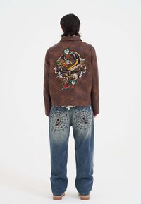 Brun läderjacka med en färgglad drake och blommig broderi på ryggen. Ljust blå denimjeans med spindelnätsmönster på fickorna.