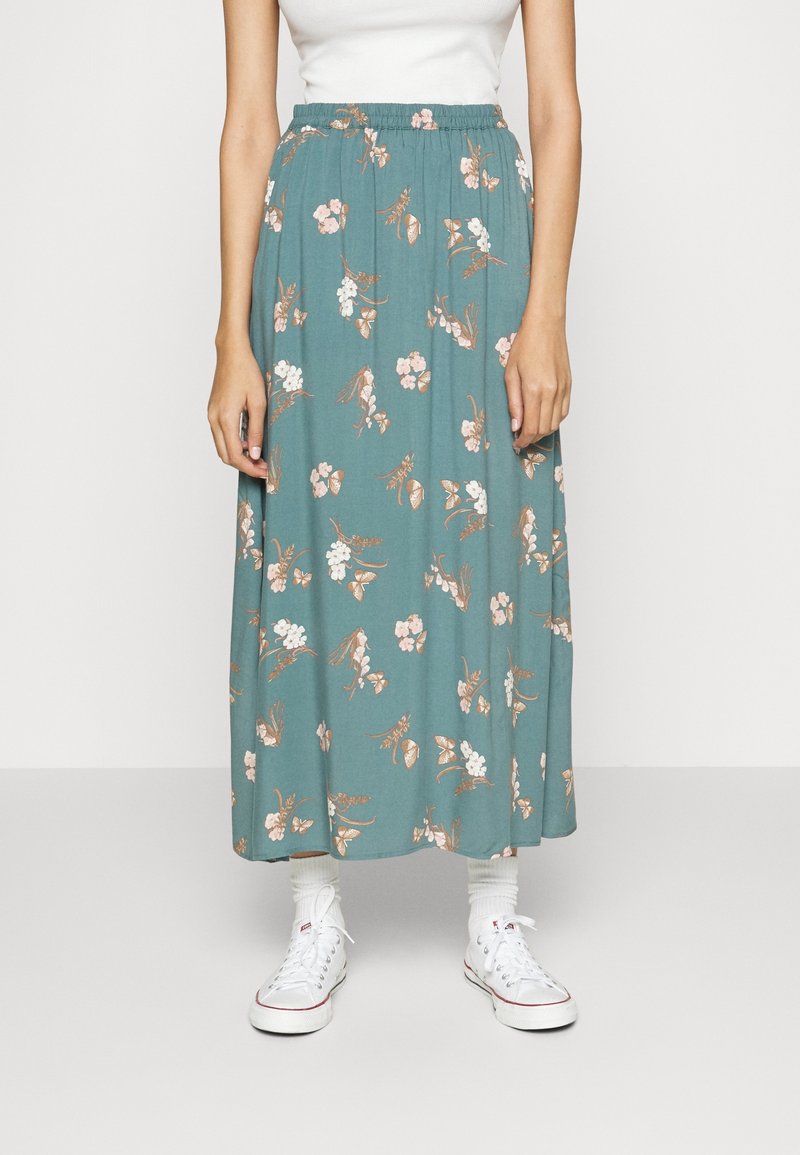 Femme portant une jupe midi florale turquoise avec une ceinture élastique, un débardeur blanc, des chaussettes blanches et des baskets blanches, debout devant un fond uni.