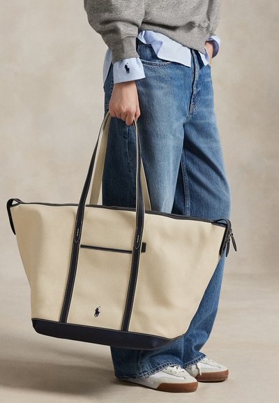 Person iført blå jeans og grå sweater holder en stor beige tote taske med sort kant og en sort logo nær bunden.