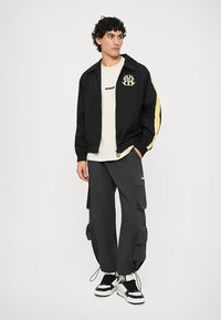 Must mustaar bomber-jakk, millel on kollased triibud ja logo rinnal, kantud valge T-särgi peal. Tumedad kaubanduslikud püksid, millel on mitmed taskud ja reguleeritavad sääred.