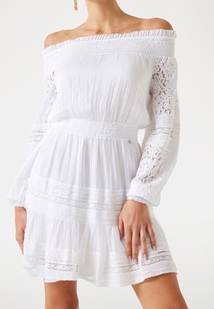 Kvinde iført hvid off-shoulder kjole med blonder lange ærmer og elastisk talje, stående med hånden hvilende på låret.