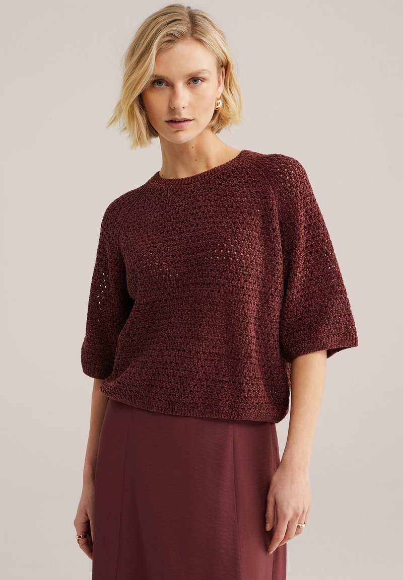 Heledate juustega naine, kes kannab tekstuuriga tume punast poolvarrukatega kootud kampsunit ja sobivat tumespunast lihtsat seelikut beeži tausta ees.