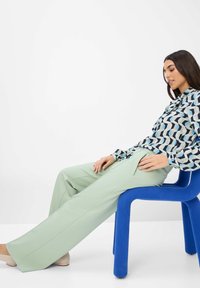 Blauer Plastikstuhl mit abgerundeten Kanten, der eine sitzende Person stützt, die mit einer gemusterten Bluse und hellgrünen, weit geschnittenen Hosen gekleidet ist.