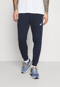 Námořnické modré tepláky s elastickými manžetami a bílým logem Nike na levém stehně, doplněné o šedé sportovní boty s oranžovými detaily.