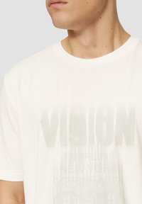 Witte katoenen T-shirt met een grafische opdruk van "VISION" en stadsnamen in lichtgrijs. Ronde hals, losse pasvorm, korte mouwen.
