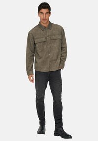 Giacca a camicia in faux suede marrone con due tasche sul petto, chiusura a bottoni, maniche lunghe, abbinata a jeans neri e stivali neri.