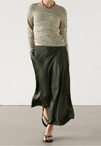 Pull en tricot beige clair avec un large col, associé à une longue jupe fluide en satin vert olive et des tongs noires.