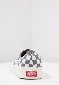 Svart-vitt rutigt Vans-sneaker med vit sula, sedd bakifrån som visar den röda "Vans Off The Wall"-logotypen på hälen.