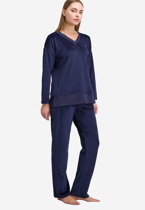 Femme debout pieds nus portant un haut à manches longues en velours bleu marine et un pantalon assorti sur un fond blanc uni.