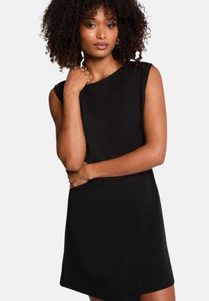 Femme aux cheveux bouclés portant une robe noire sans manches, posant avec une main près du cou et l'autre au niveau de la taille.