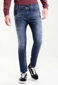 Herrjeans i blå denim med slitna detaljer, avsmalnande passform och drachordmidja. Matchas med svarta sneakers med vita inslag.