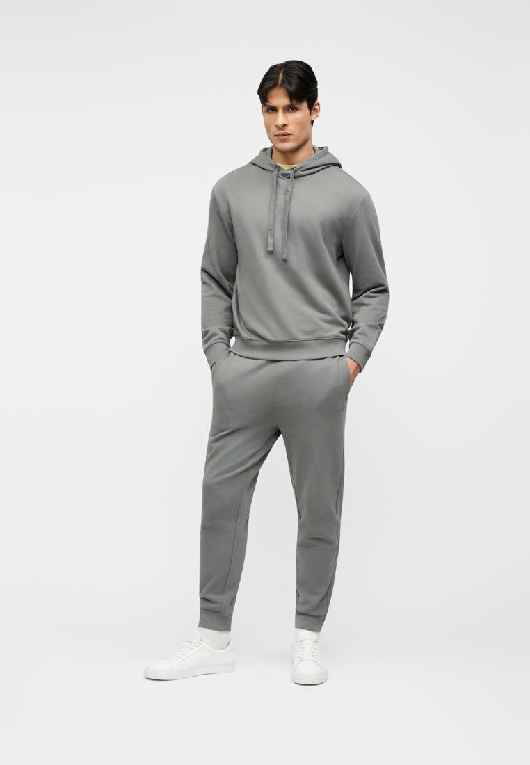 Homme debout les mains dans les poches, portant un sweat à capuche gris avec un jogging assorti et des baskets blanches, sur un fond blanc uni.