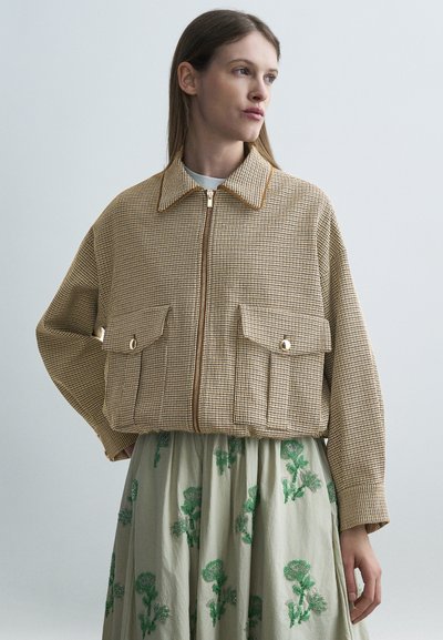 Femme aux cheveux longs et raides portant une veste beige à carreaux avec fermeture éclair et poches à rabat, associée à une jupe claire avec broderie florale verte.