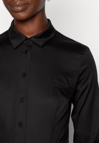 Schwarze Button-Down-Hemd aus glatter Stoffqualität mit schmalem Kragen und langen Ärmeln, ausgestattet mit einem gestickten Logo auf der Brust.