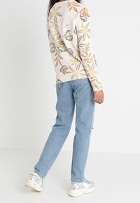 Beiger Pullover mit langen Ärmeln und buntem Blumenmuster, kombiniert mit hellblauen Jeans und silbernen Sneakers. Lässiger Schnitt, entspannter Stil.